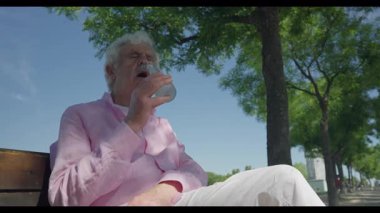 Madrid 'den emekli adam pembe gömlekli, ağaçların altındaki bankta huzur içinde oturuyor, elinde bir içki şişesi ve güneş ışığında kapalı gözlerle anın tadını çıkarıyor..