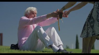 Yaşlı bir adamın çimlerden kalkmasına yardım etmek için elini uzatan bir kadın için ilgi dolu bir an. İkisi de dışarıda neşeli bir bağ paylaşıyor..