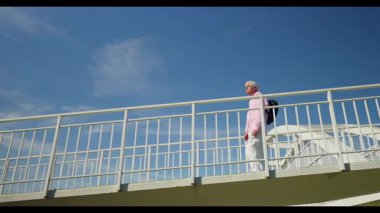 Pembe gömlekli ve beyaz pantolonlu yaşlı adam güneşli bir günde yaya köprüsünde yürürken neşe içinde dans ediyor, açık mavi gökyüzü altında hayatın ve hareketin tadını çıkarıyor..