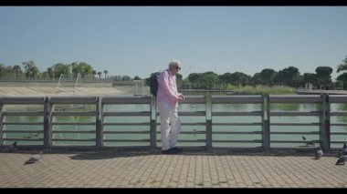 Yaşlı bir insan neşeli bir şekilde, renkli kuşlar ve doğal güzelliklerle çevrili huzurlu bir su gövdesinin yanında oturur.