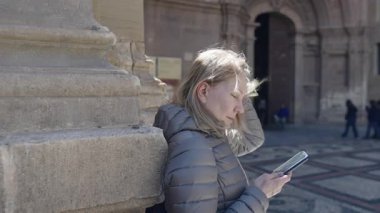 Gri ceketli sarışın kadın taş bir sütunun yanında duruyor ve telefonunu tarihi bir kilise girişinin yanında kontrol ediyor. Rüzgâr saçlarını savuruyor ve güneş ışığı sahneyi aydınlatıyor..