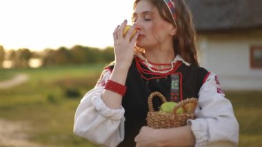 Young woman in folk costume with a black vest and traditional embroidery stands outdoors holding a yellow apple and basket, with a village house in the background.