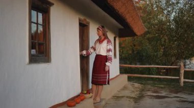 Young woman in traditional embroidered clothes stands by an old house holding a basket filled with apples and pumpkins. She looks ahead as if preparing to walk, framed by warm autumn light.
