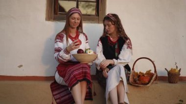 Nakışlı halk kıyafetleri içindeki genç kadınlar verandada bir sepet elma ve balkabağıyla oturuyorlar. Bir tanesi nazikçe elinde küçük bir balkabağı tutuyor..