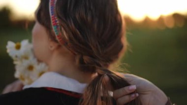 Close-up of a girl with loose wavy hair holding daisies while another pair of hands carefully braids her hair in soft evening light, creating a peaceful rural moment.