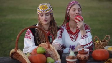Nakışlı elbiseli bir kadın dudaklarının yanında kırmızı bir elma tutarken diğeri kırsal bir masada meyve ve balkabağı dolu bir hasat sepetine bakıyor..