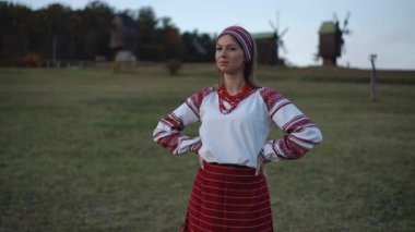 A young woman in an embroidered folk blouse and red beads stands with crossed arms in a grassy field. Her calm and confident expression highlights the grace of traditional heritage.