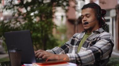 Genç bir adam dışarıda dizüstü bilgisayarıyla çalışıyor, kulaklık takıyor, doğanın güzelliği ile çevrili.