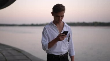 Şık bir genç adam, huzurlu ve huzurlu bir günbatımında nehir kenarında akıllı telefonuyla zarif bir şekilde etkileşime girer.