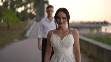 Beyaz elbiseli gelin kameraya ışıl ışıl gülümserken beyaz gömlekli damat arkasından yürüyor. Ağır çekimde yakalanan neşeli an aşkı ve mutluluğu vurgular..