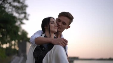 Beyaz gömlekli, zarif beyaz elbiseli gelin gün batımında suyun kenarında sevgi ve bağlılık gösteren şefkatli bir kucaklaşmayı paylaşıyor..