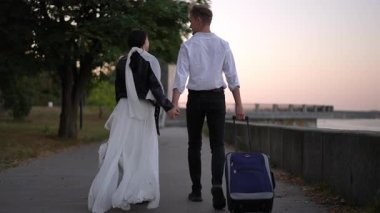 Genç çift nehir kenarındaki yolda el ele yürür, adam bavul taşır ve kadın ceketli, yeni yolculuklarının başlangıcını sembolize eder.