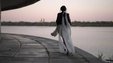 Şen elbiseli ve deri ceketli neşeli genç bir kadın gün batımında neşe içinde nehrin kenarında dönüyor. Atkısı ve elbisesi ağır çekimde rüzgarda hareket ediyor.