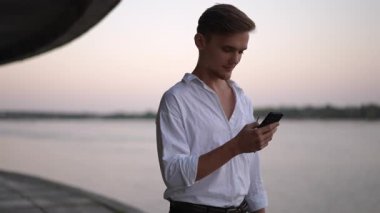 Güneş gözlüklü beyaz gömlekli bir adam gün batımında nehir kenarında akıllı telefonunu kontrol ederken gülümsüyor. Rahat ve samimi bir şekilde, açık havada düğün atmosferi..