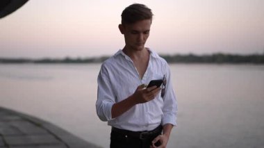 Beyaz gömlekli gülen damat gün batımında nehir kenarında duruyor. Elinde yeşil ekranlı akıllı bir telefonla el hareketi yapıyor. Açık havada eğlenceli, yavaş çekim bir düğün sahnesi..