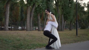 Neşeli damat, parkta yürürken beyaz elbiseli gülen gelini kaldırıyor. Sevgi ve mutlulukla dolu neşeli bir düğün anı..