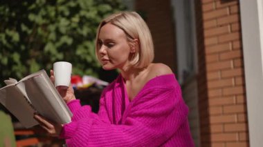 Pembe kazaklı bir kadın, güneşli bir günde kahve içerken kendini kitaba kaptırmış bir şekilde dışarıda oturuyor.