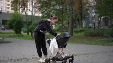 Bir kadın şık bir bebek arabasını huzurlu bir park ortamında itiyor. Güzel bir şekilde modern ebeveynlik gösteriyor.
