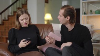 Bir kadın akıllı telefonuna hayal kırıklığıyla bakarken kocası konuşmaya çalışıyor, iletişim eksikliğini, duygusal uzaklığı ve evdeki ilişkilerinde büyüyen çatışmayı vurguluyor..