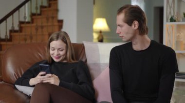 Genç bir kadın akıllı telefonuna gülümseyerek odaklanırken, kocası ona endişeyle bakıyor. Sahne, evdeki aile ilişkilerinde dikkat dağınıklığı, ihmal ve duygusal mesafeyi gösteriyor..