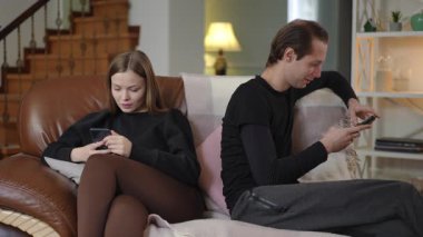 Bir erkek ve bir kadın yan yana kanepede oturur, her biri kendi akıllı telefonuna odaklanır. Sahne dijital bağımlılık, dikkat dağınıklığı ve evdeki ilişkilerinde gerçek iletişim eksikliğini gösteriyor..
