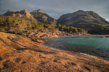 Sunlit Honeymoon Bay in Tasmania, Australia, with rugged mountains, turquoise waters, and red rocky coastline. High quality photo clipart