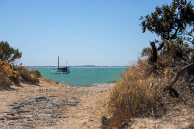 Turkuaz sularda demirli tekne Avustralya 'daki kuru çalılar ve rüzgar şeklinde bitki örtüsüyle çerçevelenmiş kumlu kıyı yolundan görülüyor.