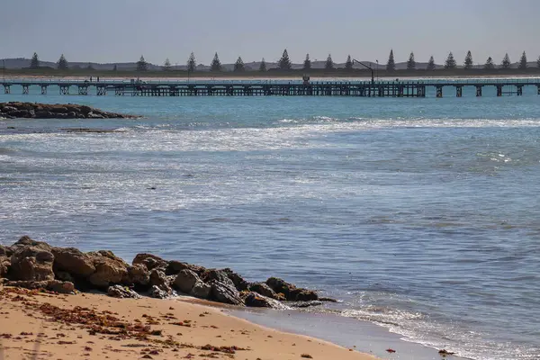 Yumuşak dalgalı güneşli kıyı manzarası, kumlu sahil, kayalık kıyı şeridi ve sakin mavi deniz boyunca uzanan uzun balıkçı iskelesi.