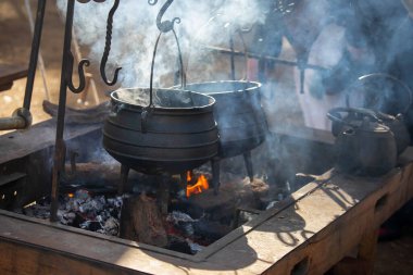 Geleneksel dökme demir tencere ve su ısıtıcısı açık ateş çukurunda asılı ve tarihi kampta yükselen dumanla birlikte.