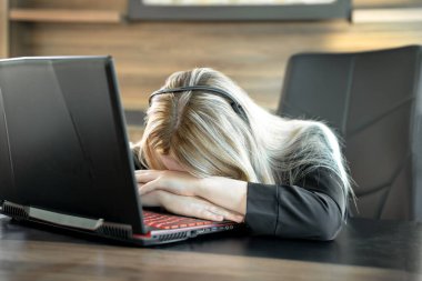 Siyah takım elbiseli yorgun genç bir iş kadını ofiste dizüstü bilgisayarın önünde oturuyor, kafası ellerinin üzerinde dinleniyor.