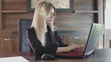 Genç sarışın kadın dizüstü bilgisayarın önünde oturuyor ve ofiste yazı yazıyor.