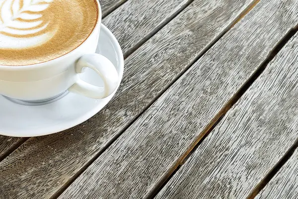 Coffee in white cup with pattern on top of foam, against background of wooden table, copy space