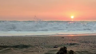 Açık deniz dalgaları kıyıya vurur pembe günbatımının arka planında beyaz köpüğe dönüşür.