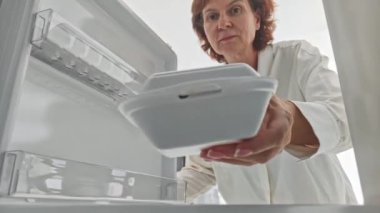 Orta yaşlı bir kadın dondurucuyu açıyor ve yiyecekleri tek kullanımlık beyaz konteynıra koyuyor..