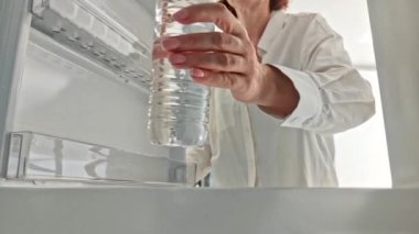 Kadın soğutmak için buzdolabına küçük plastik bir şişe su koyuyor. Buzdolabının içini görüyor..