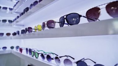 Beyaz raflarda sergilenen renkli güneş gözlükleri, gözlük seçimleri ve moda aksesuarları, perakende deneyim ve canlı alışveriş ortamı yaratıyor. .