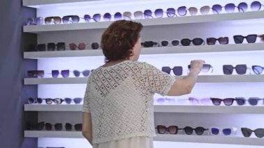 Kadın, çeşitli stillerle çevrili güneş gözlüğü koleksiyonunu inceliyor, tasarım seçeneklerini araştırıyor, tercih edilen görünüm, çağdaş iç mekan, gözlük seçimi, moda alışveriş deneyimi