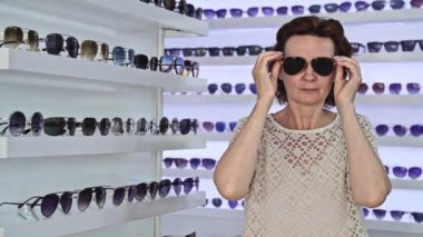 Güneş gözlüğü deneyen bir kadın, şık çerçevelerle çevrili, modern perakende seti, gözlük alışverişi, kişisel stili keşfetmek, göz gezdirmek, görünüşü değerlendirmek, moda deneyiminden zevk almak.