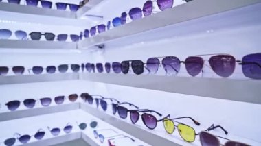 Beyaz raflarda sıralanmış güneş gözlükleri, çok renkli lens renkleri ve ileri moda stilleri, organize perakende sunumu ve davetkar bir alışveriş ortamı yaratıyor.