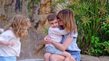 Güneş gözlüklü anne güneşli bir günde egzotik parkta şelaleyle oturuyor, oğluna sarılıyor, kameraya gülümsüyor, sarı kıvırcık saçlı kız ise neşe dolu aile ortamında oynaşıyor..