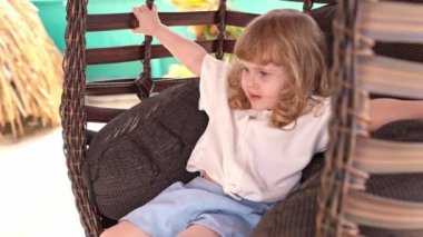 Küçük kıvırcık sarışın kız rahat bir şekilde salıncakta oturuyor, rahat bir duruş sergiliyor, sakin bir anın tadını çıkarıyor, egzotik bir arka plan, boş zaman, tasasız bir çocukluk, rahat bir ortam,