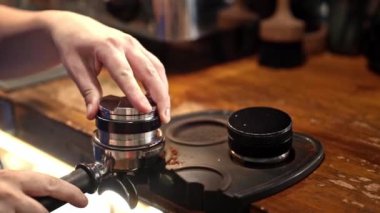 Barista titizlikle öğütülmüş kahveye basıyor, espresso hazırlıyor, ustalığa odaklanıyor, detaylara bağlılığı sembolize ediyor, ve sıcak bir kafe ortamında kahve yapma sanatını simgeliyor..