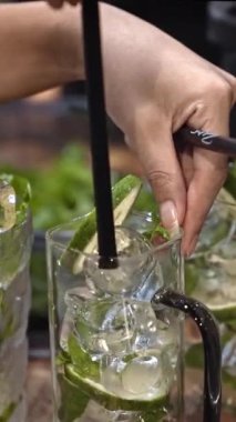 Barmen bardağa saman koyar, limonlu ve naneli el yapımı kokteyl, ferahlatıcı içecek hazırlığı, berrak bardakta taze malzemeler, yazın sembolü, serinletici ferahlatıcı, dikey.