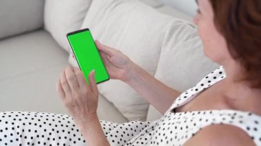 Orta Yaşlı Kadın Yeşil Ekran Chromakey ile Akıllı Telefon Kullanıyor Koltukta oturan, Dokunmatik Ekranla Yakınlaşan, Modern Teknoloji Konseptiyle Doğal Gün Işığında Rahat Ev Ayarları.
