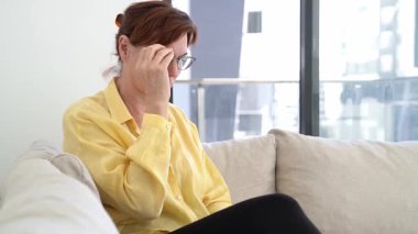 Rahat bir yuva ortamında, sarı gömlekli olgun bir kadın gözlüğünü takar ve yumuşak bir koltukta otururken akıllı telefonuyla meşgul olur, odaklandığını ve rahatladığını gösterir..