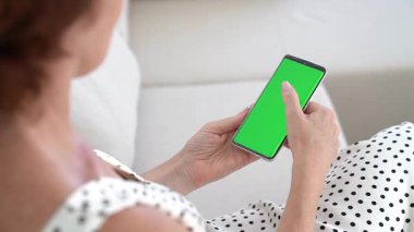 Kanepede rahat esmer bir kadın, elinde akıllı telefon, yeşil renkli ekran aydınlatmalı, rahat ev düzeni, dijital etkileşim, çağdaş yaşam tarzı, kablosuz iletişim..