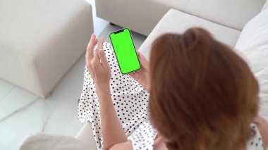 Esmer kadın koltukta rahat, akıllı telefon yeşil ekran sergiliyor, modern ev dekorasyonu, dijital iletişim, çağdaş yaşam tarzı, kablosuz teknoloji, kucaklama, üst görünüm.