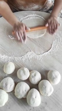 Sıcak bir mutfakta, bir kadın ahşap bir pim kullanarak ustalıkla hamuru çevirir, elleri un tozuyla kaplıdır. Masa düzenli hamur toplarıyla süslenmiş, geleneksel gözleme hazırlama sanatını gözler önüne seriyor..