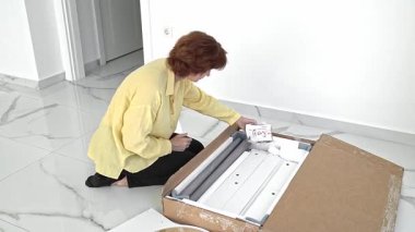 Bir kadın bir kutu mobilyayı dikkatlice açıyor, montaj için gerekli tüm parça ve aksesuarları kontrol ediyor. Yeni mekanı ve yeni ev hazırlıkları için heyecanlandığını gösteriyor..