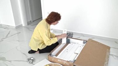 Bir kadın hevesle mobilya parçalarıyla dolu bir kutuyu açıyor, özenle parçaları kontrol ediyor ve montaja hazırlanıyor. Aydınlık alan, yaklaşan ev düzenine heyecan katıyor..